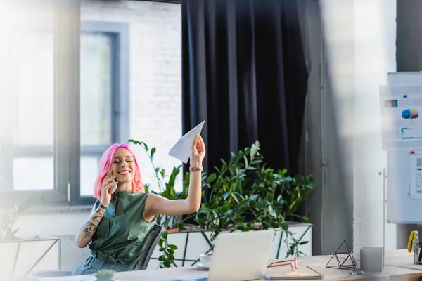 Positive businesswoman with pink hair talking on smartphone while throwing paper plane — Stock Photo