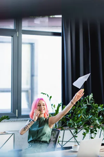 Manager positivo con i capelli rosa che parla sullo smartphone mentre lancia aereo di carta — Foto stock