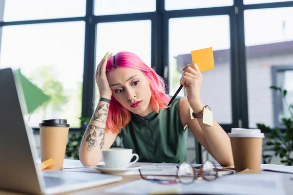 Gerente cansado con el pelo rosa celebración de pluma cerca de tazas de papel con café en la oficina - foto de stock