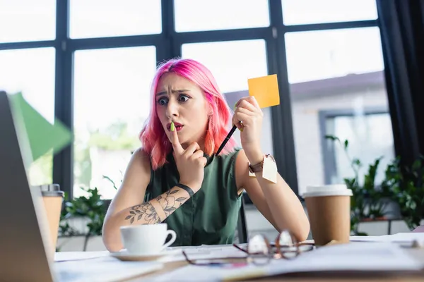 Shocked manager with pink hair holding pen near paper cups with coffee in office — Stock Photo