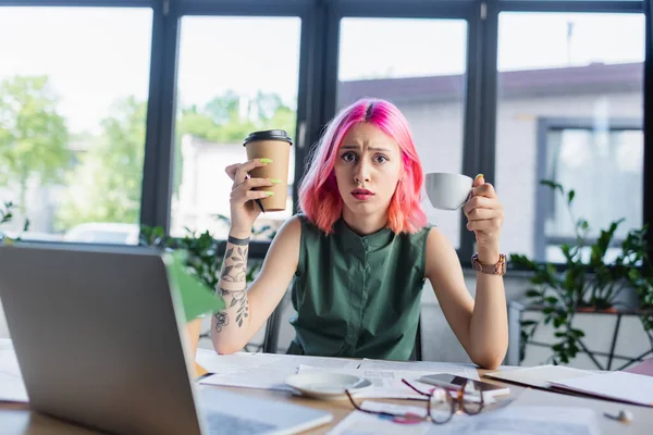 Шокований менеджер з рожевим волоссям тримає каву біля пристроїв в офісі — стокове фото