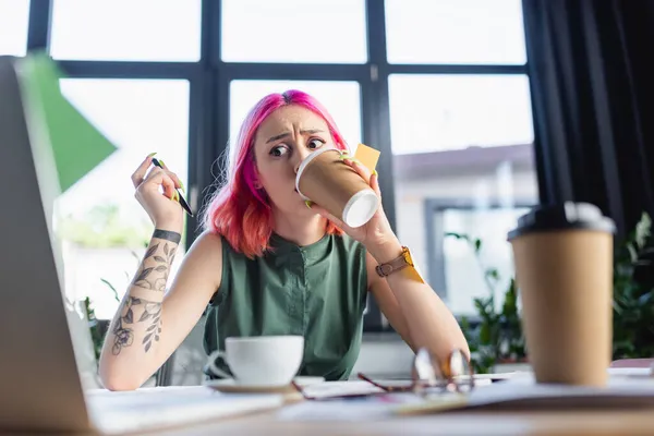 Femme d'affaires choquée avec les cheveux roses boire du café près d'un ordinateur portable au bureau — Photo de stock