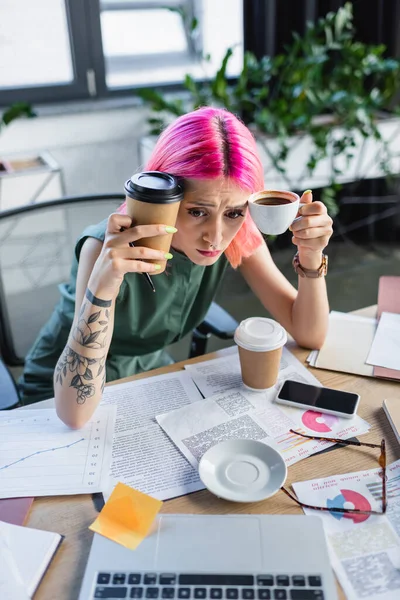 Vista de ángulo alto de la mujer de negocios estresada con cabello rosa sosteniendo café y mirando a la computadora portátil - foto de stock