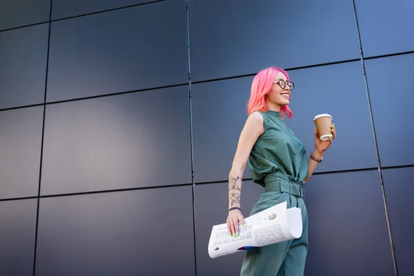 Счастливая и татуированная деловая женщина с розовыми волосами, держащая бумажную чашку и графики снаружи — стоковое фото