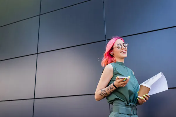 Femme d'affaires heureuse et tatouée dans un casque tenant smartphone et tasse en papier — Photo de stock