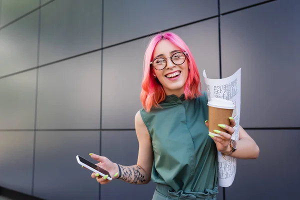 Felice donna d'affari con capelli rosa e auricolare in possesso di smartphone e tazza di carta — Foto stock