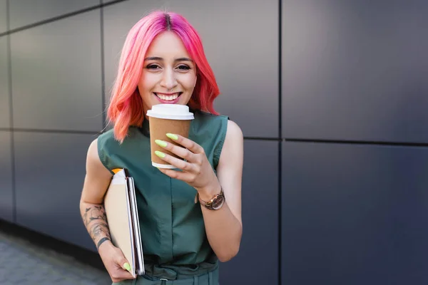 Feliz mujer de negocios con el pelo rosa y auricular sosteniendo café para ir y carpetas - foto de stock