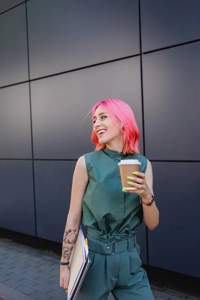 Mulher de negócios sorridente com cabelo rosa e fone de ouvido segurando café para ir e pastas — Fotografia de Stock