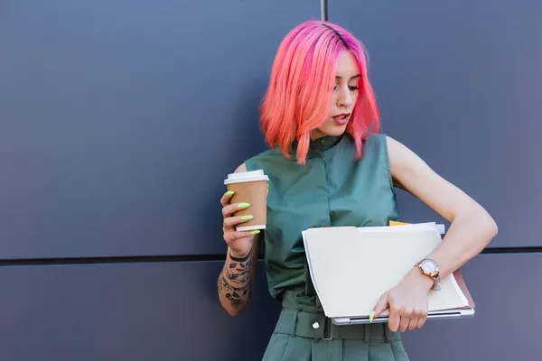 Tätowierte Geschäftsfrau mit rosa Haaren, die Ordner, Laptop und Kaffee nach draußen hält — Stockfoto