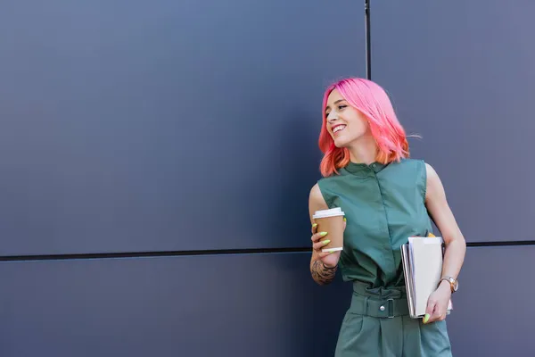 Татуированная и улыбающаяся деловая женщина с розовыми волосами, держащая папку и кофе, чтобы выйти наружу — стоковое фото
