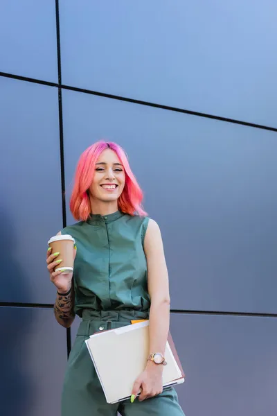 Tätowierte und positive Geschäftsfrau mit rosa Haaren, die Ordner, Laptop und Kaffee nach draußen hält — Stockfoto