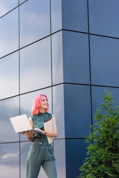 Femme d'affaires tatouée et joyeuse avec des cheveux roses tenant ordinateur portable et dossier à l'extérieur — Photo de stock