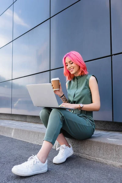 Mulher de negócios positiva com cabelo rosa segurando copo de papel e usando laptop fora — Fotografia de Stock