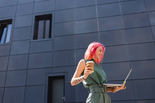 Feliz mujer de negocios con el pelo rosa sosteniendo taza de papel y el uso de ordenador portátil fuera - foto de stock