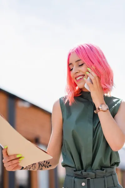 Geschäftsfrau mit pinkfarbenen Haaren hält Ordner in der Hand und telefoniert draußen auf dem Smartphone — Stockfoto