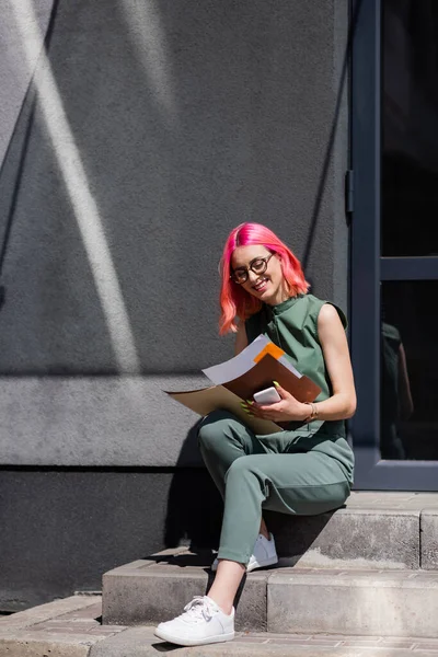 Donna d'affari soddisfatto con capelli rosa cartella con documenti e smartphone mentre seduto sulle scale fuori — Foto stock