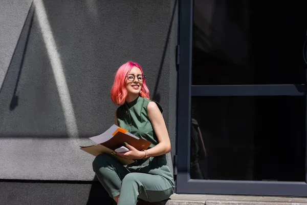 Glückliche Geschäftsfrau mit rosafarbenen Haaren hält Ordner mit Dokumenten und Smartphone in der Hand, während sie draußen auf der Treppe sitzt — Stockfoto