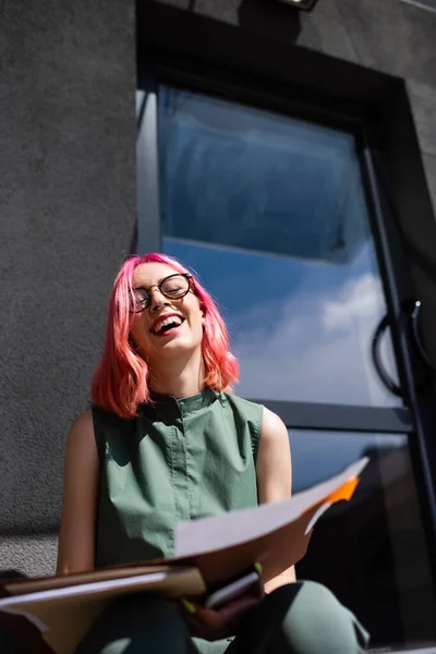 Visão de baixo ângulo de empresária alegre com cabelo rosa segurando pasta e smartphone enquanto sentado fora — Fotografia de Stock