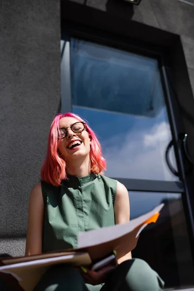 Vista a basso angolo di gioiosa donna d'affari con cartella di tenuta dei capelli rosa e smartphone mentre si siede fuori — Foto stock