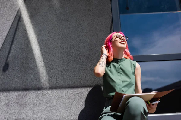 Mujer de negocios complacida con carpeta de retención de pelo rosa con documentos y teléfono celular mientras está sentada afuera - foto de stock