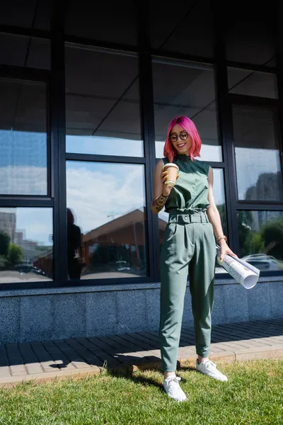 Full length of pleased businesswoman with pink hair holding rolled paper and coffee to go outside — Stock Photo