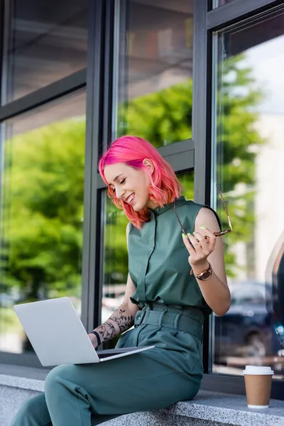 Allegra donna d'affari con capelli rosa che tiene gli occhiali e utilizza il computer portatile all'esterno — Foto stock
