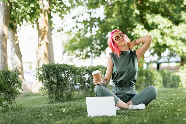 Mulher satisfeita com cabelo rosa e óculos segurando copo de papel perto de laptop na grama — Fotografia de Stock