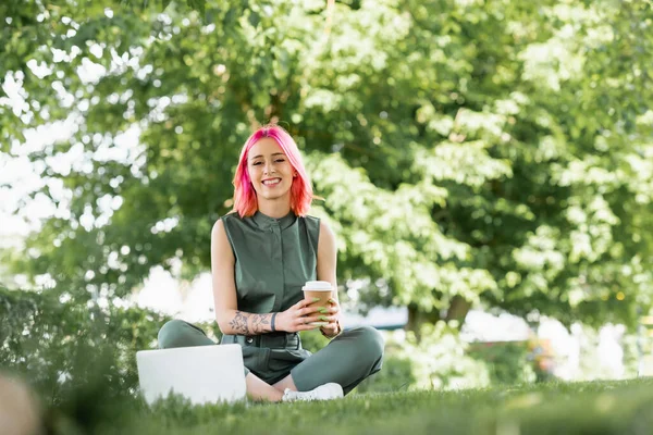 Heureuse jeune femme avec les cheveux roses tenant tasse en papier près de l'ordinateur portable sur l'herbe — Photo de stock