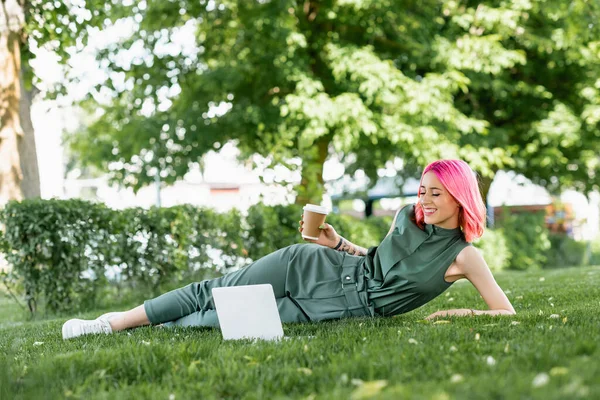 Усміхнена молода жінка з рожевим волоссям тримає паперову чашку біля ноутбука на траві — стокове фото