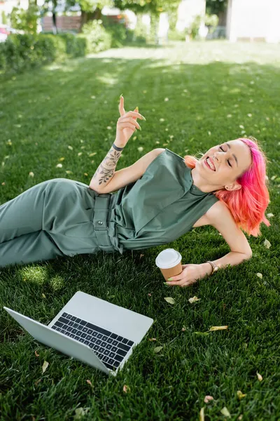 Mujer feliz con el pelo rosa y los ojos cerrados sosteniendo taza de papel cerca de la computadora portátil en la hierba - foto de stock