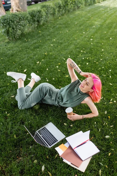 Vue grand angle de femme heureuse avec les cheveux roses et les yeux fermés tenant tasse en papier près de l'ordinateur portable sur l'herbe — Photo de stock