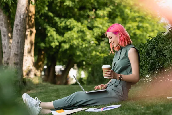 Вид сбоку счастливой женщины с розовыми волосами, держащей бумажный стаканчик и использующей ноутбук на траве — стоковое фото
