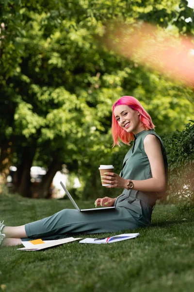 Mulher feliz com cabelo rosa segurando copo de papel e usando laptop na grama — Fotografia de Stock