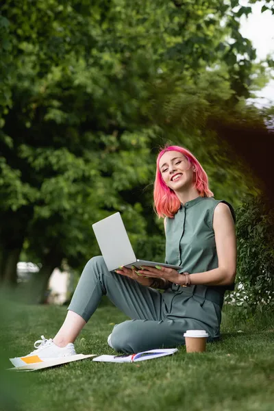 Donna gioiosa con i capelli rosa che tiene il computer portatile e seduto sull'erba — Foto stock