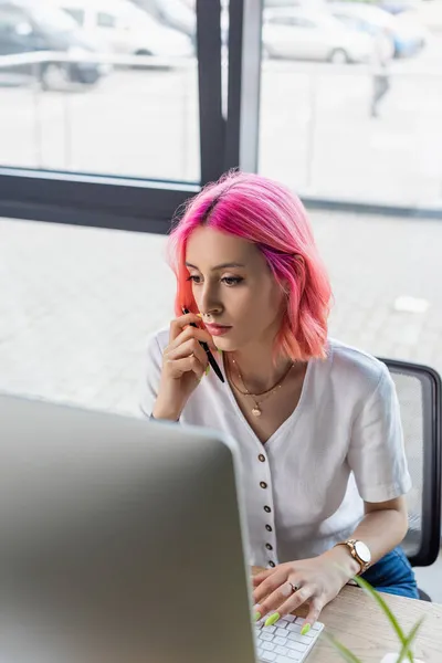 Femme d'affaires concentrée avec des cheveux roses tenant stylo près de l'ordinateur moniteur — Photo de stock