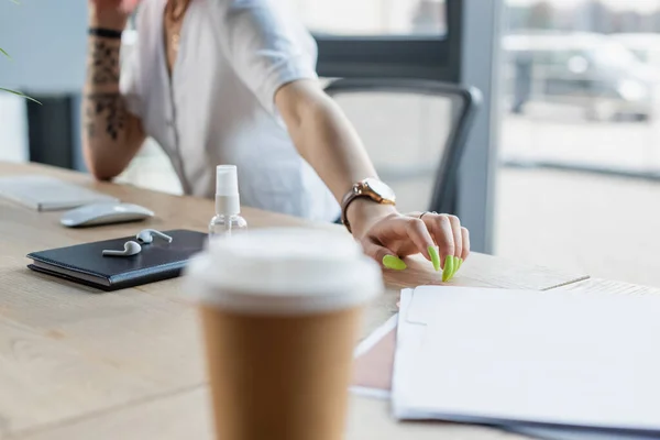 Vista recortada de mujer de negocios tatuada cerca de taza de papel borrosa en primer plano - foto de stock