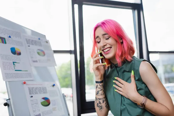 Femme gaie et tatouée avec les cheveux roses parler sur smartphone au bureau — Photo de stock