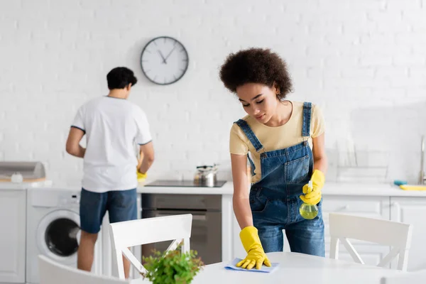 Africana americana mujer limpieza mesa de comedor con trapo cerca borrosa novio - foto de stock