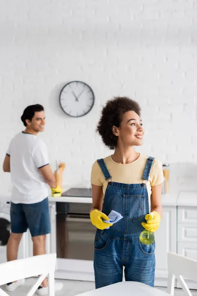 Heureuse femme afro-américaine tenant vaporisateur bouteille et chiffon près de petit ami flou — Photo de stock