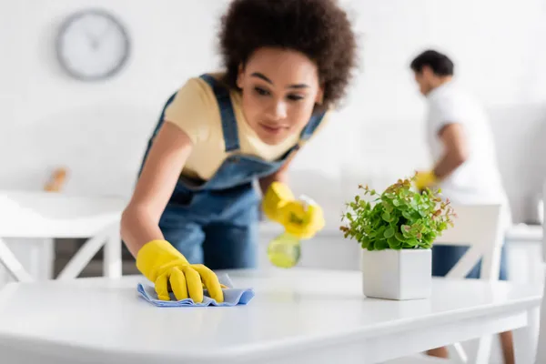 Borrosa africana americana mujer limpieza mesa de comedor con trapo cerca borrosa novio - foto de stock