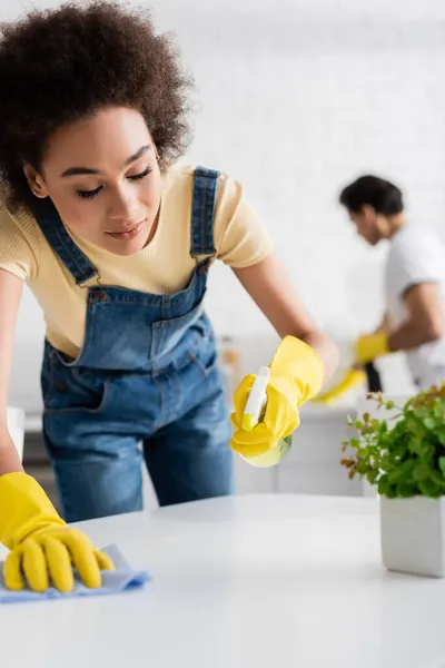 Encaracolado afro-americano mulher limpeza mesa de jantar com pano perto de homem borrado — Fotografia de Stock