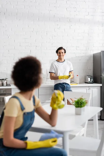 Fröhlicher Mann mit Schwamm und Blick auf verschwommene afrikanisch-amerikanische Frau — Stockfoto