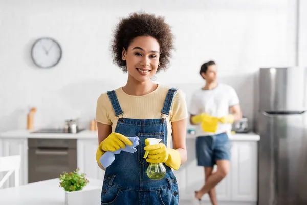 Allegra donna afro-americana con bottiglia spray e straccio vicino all'uomo sfocato in cucina — Foto stock