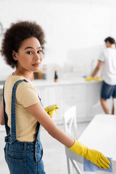 Encaracolado afro-americano mulher segurando spray garrafa e pano perto da cadeira e homem borrado na cozinha — Fotografia de Stock