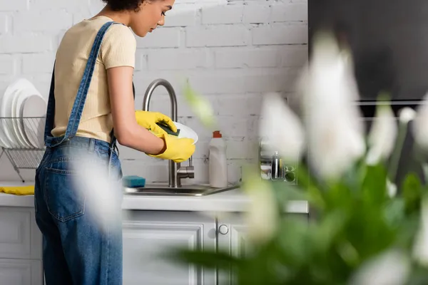Mujer joven afroamericana en guantes de goma lavando platos en la cocina - foto de stock