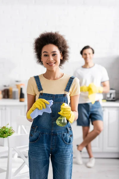 Femme afro-américaine souriante tenant vaporisateur et chiffon près de l'homme flou dans la cuisine — Photo de stock