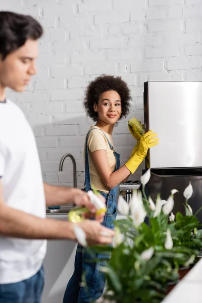Donna afro-americana sorridente che tiene la spugna vicino al frigorifero vicino all'uomo offuscato che innaffia le piante in cucina — Foto stock