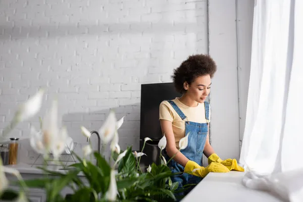 Curly african american woman holding rag near window sill and plants in kitchen — Stock Photo