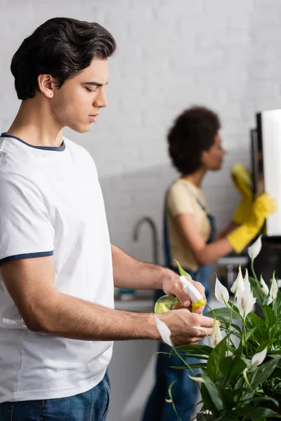 Joven hombre rociando agua en planta cerca borrosa africana americana mujer - foto de stock