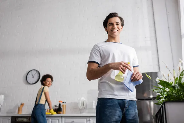 Vista de ángulo bajo de hombre joven feliz sosteniendo botella de spray y trapo cerca de plantas y borrosa mujer afroamericana - foto de stock